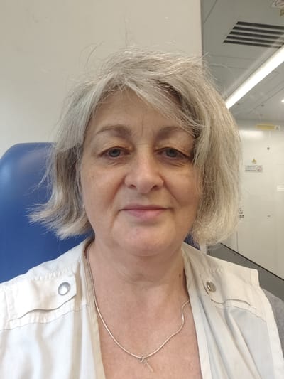 A medium close-up, eye-level selfie shows a fair-skinned, middle-aged white woman with short, graying blonde hair and blue eyes. She is looking directly at the camera with a neutral expression. She is wearing a white collared shirt with a silver button and a thin silver chain with a cross pendant. The background is slightly out of focus, showing a blue chair to her left and a wall with some fluorescent lighting and what appears to be a ceiling vent to her right. There are no strong indications of time of day or weather, but the indoor lighting suggests it is daytime. The presence of a uniform chair and the wall details may suggest she is in a public building, possibly a waiting area. No text is clearly visible or decipherable in the image.