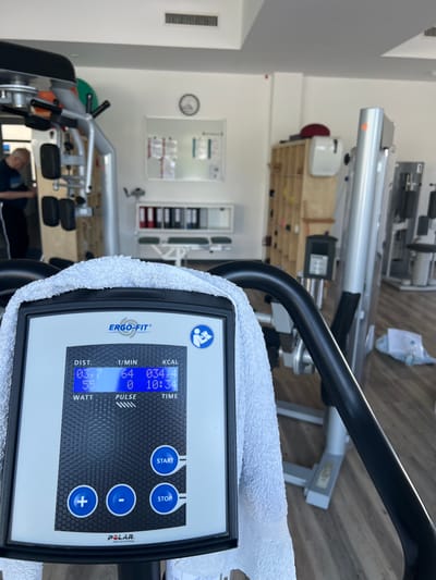 The image shows a close-up of a fitness machine console with a white towel draped over it. The console displays workout statistics, including distance, speed, calories burned, and heart rate. The brand "Ergo-Fit" is visible on the console, along with "Polar heart rate technology". In the background, the interior of a gym or rehabilitation center is visible, with other exercise equipment and storage shelves. A person in workout attire is partially visible in the left background, appearing to be using another machine. The environment appears to be well-lit and clean, with wooden floors. The scene is devoid of any explicit clues identifying it as being located in Teltow, Germany.