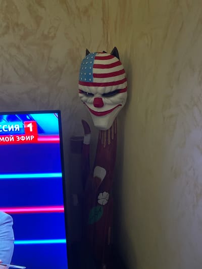A still indoor shot shows a wooden cat figurine painted with the American flag colors, lying sideways on a surface. The head is painted to resemble the American flag, while the body is brown with a white flower and green leaf design. The figurine is on top of a piece of furniture. In the foreground, the bottom of a TV screen displays a program in Russian, with a logo including the number "1" and Cyrillic text. The image was taken in Лесной Пейзаж Кп (шарапово), Russia.