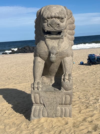A weathered grey stone statue of a Foo Dog (Chinese Guardian Lion) stands centrally on a light tan sandy beach in Long Branch, United States. The statue faces forward, featuring an open mouth with a visible spherical object inside, and its left front paw rests on another spherical object. A stylized bell-like ornament is suspended from its chest. The statue is positioned on a carved rectangular stone base adorned with floral and geometric patterns.

Behind the statue, the dark blue ocean exhibits white breaking waves. A dark rock jetty, composed of large boulders, extends into the water on the left side of the frame. The sky above is clear and light blue. To the right of the statue, further back on the sand, a dark beach wagon containing various items and a small blue spherical object are present. Strong sunlight illuminates the scene, casting a distinct shadow of the statue onto the sand to the viewer's right. No individuals or discernible actions are visible.