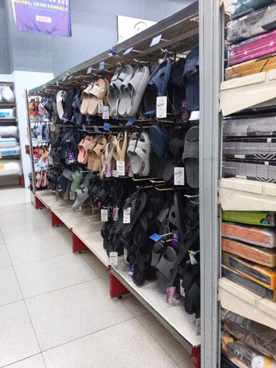 Aisle in a retail store featuring a large selection of sandals and flip-flops. The footwear hangs from hooks along a shelving unit. A variety of colors and styles are visible, mainly in neutral tones like beige, gray, and black. The opposite shelf showcases stacks of packaged goods, possibly textiles or home goods, in colorful layers. A banner with promotional text is visible above. The floor is tiled and the overall lighting is bright.