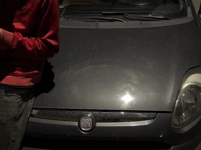 The image shows the front of a dark-colored Fiat car, possibly a Grande Punto or similar model, in what appears to be an outdoor setting. The car's hood is visibly dirty with some circular marks, possibly from cleaning. The front of a person, wearing a red sweatshirt and dark jeans, is partially visible on the left side of the frame, leaning against the vehicle. The background is not clearly visible, but based on the image, it is not possible to confirm the specific location as Amarante, Portugal. No specific actions or interactions are observable beyond the person's proximity to the vehicle. The environment is dimly lit.