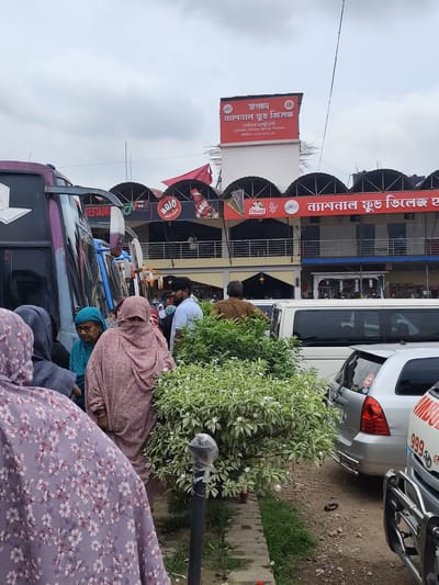 The image captures a scene at the National Food Village in Hatikumrul, Bangladesh. A group of people, some wearing traditional head coverings and floral-patterned clothing, are gathered near buses and cars. There are multiple buses parked on the left. Among the vehicles is an ambulance with the number 999 on the side. A bush with white flowers is visible in the foreground. The National Food Village building has signs with its name in both English and Bengali, along with advertisements for various food items and beverages such as Mojo. The sky is overcast.