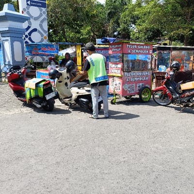 The scene takes place in the city of Kendal, Indonesia, at a location with coordinates -6.921 latitude and 110.204 longitude. There are three motorcycles parked on a paved surface. One motorcycle is red and black, another is white and black, and the third is white and grey. A man in a green and yellow reflective vest stands next to the white and grey motorcycle and faces towards a red and white food cart. The food cart has a sign that reads "Telur Gulung & Sempolam" in red lettering with black and white graphics of food. The man is looking at the food cart. A woman wearing a red and black hijab is sitting on a stool on the other side of the food cart. She is looking at the red and black motorcycle. The food cart has a large window that displays food. There is also a white sign next to the food cart. The sign reads "Telur Gulung & Sempolan" in red lettering with black and white graphics of food. There are two other food carts that are obscured in the background. There are trees in the background. The scene is well lit by the sun. The surface of the ground is grey. There is a blue and white structure with the word "Galoei ATM" on it in the top left of the frame. There is a yellow and white sign on the ground that reads "Pasa Godok Bakar" in black lettering with a small image of a grill. The scene is quiet and serene. There is no evidence of any traffic or other activity.