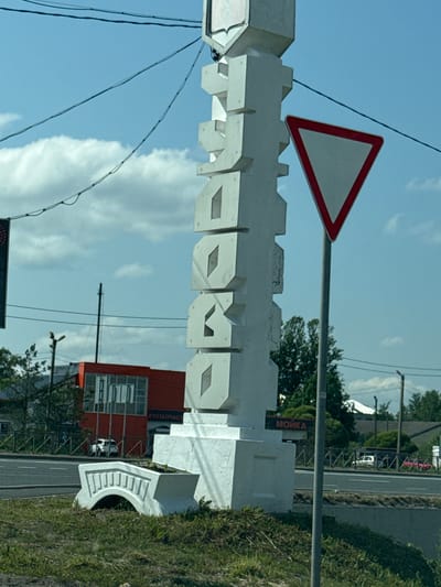 This image captures a street scene in Chudovo, Russia. In the foreground, a large, white, abstract sculpture stands prominently, featuring stacked, geometric blocks with cutouts. To the right of the sculpture is a triangular "Give Way" road sign on a grey pole. Behind the sculpture, a red building with large windows is visible, and to its right, a sign reads "МОЙКА" (Moyka), indicating a car wash. A white car is parked in front of the red building, and other vehicles are visible on the road. The scene is set against a backdrop of a blue sky with scattered clouds, green trees, and power lines. A small, white, decorative bridge structure is also present in the lower left foreground.