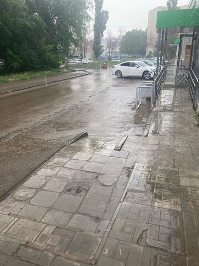 The image depicts a wet, paved area in Balakovo, Russia, under overcast skies with visible rain. A white sedan is parked on the right side of the frame, adjacent to a building with a green awning and a pharmacy cross emblem. In the background, other cars are visible on a wet road, along with trees and buildings. The foreground features a damaged, wet tiled sidewalk with puddles. A metal bicycle rack is also present on the sidewalk. The overall scene suggests a common urban environment experiencing inclement weather.