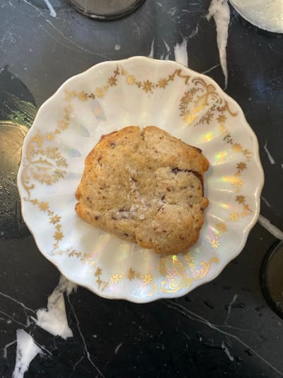 A single, light brown cookie, speckled with dark inclusions and topped with coarse white flakes, rests centrally on a small, white, iridescent plate. The plate features a scalloped rim adorned with gold-colored floral motifs and scrollwork. This plate is positioned on a polished dark surface, consistent with black marble exhibiting distinct white veining. The scene is devoid of individuals, actions, or interactions, and is located in the city of La Tour-d'Aigues, France.