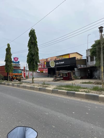 This is a street-level photograph taken in the city of Dalam, Indonesia, with a cloudy sky overhead. In the foreground, a grey asphalt road is visible, with a concrete curb and patchy grass on the side. A portion of a motorcycle's rearview mirror and the rider's legs are reflected in the lower part of the image.

Dominating the mid-ground is a row of buildings. On the left, an orange truck with a large cargo container is parked. Above the truck, a sign with the text "ME Gacoan" in pink and blue lettering is prominently displayed. Next to it are two tall, slender, dark green trees.

Further to the right, a black building with "plus kopi" in white lettering is visible, suggesting it is a coffee shop. There are other buildings with colorful signage behind and to the side of this coffee shop, indicating a commercial area. A small red kiosk with a shaded canopy is situated in front of one of the buildings. Various signs with Indonesian text are present on the buildings.

In the background, power lines are strung across the sky, supported by utility poles. A street lamp with an ornate design is also visible on the right side of the image. The overall scene depicts a typical urban streetscape in Indonesia with businesses and traffic.