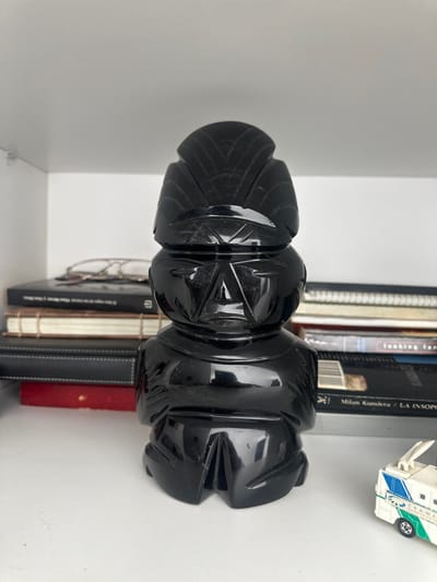 A shiny, black, ornate object sits horizontally among books in a white shelf. The object seems to be made of stacked, carved pieces. Its design features angular cuts. A toy van sits on the shelf above the object. Books with dark spines and light pages are visible behind the object, and several bound notebooks lie underneath it.