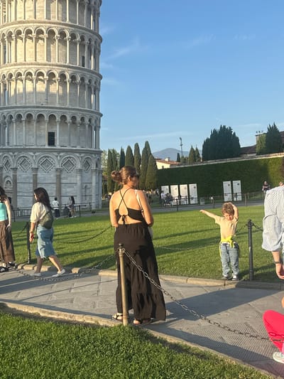 A bright, sunny outdoor photograph captures a bustling tourist scene at the Leaning Tower of Pisa, Italy. The image is presented rotated 90 degrees clockwise from its natural vertical orientation.

In the foreground, a woman with her back to the camera, wearing a black backless dress or top with criss-cross straps, stands looking towards the iconic tower. To her right, a young child, dressed in a light-colored shirt and jeans, lies on the manicured green grass, arms outstretched, seemingly engaging in the classic tourist pose of "pushing" or "holding up" the Leaning Tower.

The magnificent Leaning Tower of Pisa stands prominently on the left side of the frame, its detailed white marble architecture and characteristic tilt clearly visible under the clear blue sky. The tower's multiple levels of arched colonnades are well-defined. A paved pathway runs alongside the grassy area where people are gathered, separated by a chain barrier. Several other tourists are scattered throughout the scene; some are partially visible, including individuals in a striped shirt and red pants. Long shadows stretching across the paved path indicate that it is likely late afternoon or early evening. Lush green trees line the background, adding to the picturesque setting. While information boards are present in the mid-ground, any text on them is too small to be legible. The atmosphere is relaxed and captures typical tourist activity around this famous landmark.