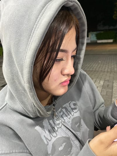 The image shows a close-up of a young woman wearing a gray hooded sweatshirt. She is looking down and to the right, with her head tilted slightly. Her hair is dark and appears to be wet or styled with product. She has a pimple on her cheek. The background is blurred, but appears to be an outdoor setting with paved ground and some lights. There are buildings and vegetation visible in the distance. The overall lighting suggests it is evening or nighttime. This scene is consistent with the environment found in Mekar Bakti, Indonesia, an area known for its residential and commercial zones.