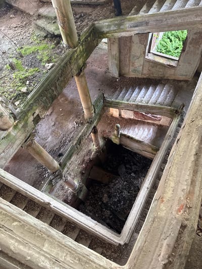 The image depicts an interior view looking downwards into a decaying, multi-level structure. The location is Tsanbaikwara, Abkhazia. The structure features weathered, crumbling concrete or plaster elements, exposed support columns, and fragmented staircases. Debris and vegetation, including moss and ferns, are present on the surfaces. A window opening reveals green foliage outside. No people are visible. The scene conveys a sense of abandonment and decay.