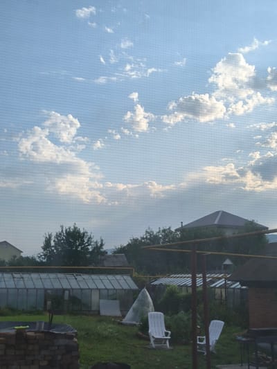 The image depicts a view of a rural or suburban setting, likely in or around Miass, Russia, given the provided context. The foreground shows a brick pile and grass with two white plastic lounge chairs. A large greenhouse structure with glass panels is visible behind the chairs, partially obscured by foliage. Further back, a house with a dark-colored roof is seen amongst trees, and a tall, thin structure, possibly a chimney or antenna, protrudes from its roof. The sky is a clear blue with scattered white clouds, suggesting daytime. The overall impression is that of a residential backyard or a dacha (summer house) property. There are no people or animals visible in the image. The scene appears to be a still, ordinary day.
