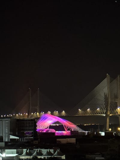 This forensic image captures a nighttime scene in the city of Loures, Portugal. The prominent feature is the Vasco da Gama Bridge, a cable-stayed structure spanning the upper and middle portions of the frame. Its multiple white towers are visible, with numerous support cables illuminated in white and connecting to the bridge deck, which is lined with small, bright yellow lights.
In the midground, directly beneath the bridge, stands a large, open-framework structure brightly illuminated in shifting hues of pink, purple, and blue, suggesting an event stage or architectural installation. To the left and below this structure are various white, peaked tents and other utilitarian buildings, consistent with event infrastructure or temporary setups.
A dark expanse of water is visible between these foreground structures and a distant landmass on the horizon, which is marked by a faint, continuous line of city lights. The sky above is dark and clear. No discernible individuals or specific actions are present in the frame.