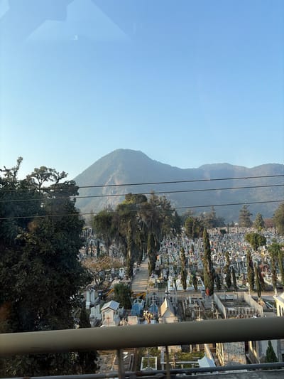 The image shows a landscape with a mountain range on the left side under a clear, blue sky. A power line runs across the mountain. On the right, a dense, grid-like layout of tombstones and structures forms a cemetery. Trees and vegetation dot the landscape between the mountain and the cemetery. A vertical metal pole is in the far right foreground, partially obscuring the view. Branches and foliage are visible at the bottom.