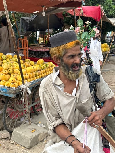 A man with blond-dyed hair and a beard, wearing a dark skullcap and a light-colored shirt, sits in front of a fruit stall in Karachi, Pakistan. He is holding a pink and white string in his hands. To his left, a cart is piled high with yellow mangoes. In the background, other market stalls and people are visible. The scene is outdoors, under a makeshift canopy.