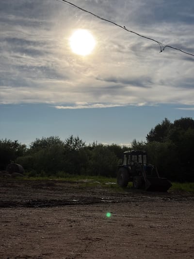 The image captures an outdoor, rural, or semi-rural scene, likely in Chumna, Russia, oriented with the sky on the left and the ground on the right. When viewed correctly, the upper portion of the frame features a bright, intense sun high in the sky, casting a strong glare. The sky is a mix of clear blue and wispy, scattered clouds.
A power line with small attachments or insulators stretches diagonally across the upper left quadrant of the sky. Below the horizon line formed by the sky, a dense line of dark green trees and foliage extends across the middle of the image. On the right side, a dirt or gravel road occupies the foreground and midground, with some green grass and vegetation bordering it. A dark-colored tractor or heavy machinery, possibly a front-end loader, is partially visible behind the road and amidst the trees on the left side. A prominent bright green lens flare is visible on the dirt road, indicating the direct sunlight.
The scene depicts a bright, clear day, suggesting morning or afternoon given the sun's position. No people or specific activities are visible. There is no visible text in the image.