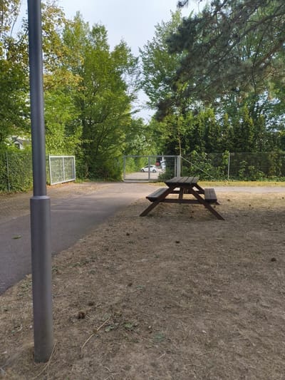 A metal lamppost stands in the foreground, partially obscuring a paved path. Beyond the path, a wooden picnic table with benches is situated on dry, patchy grass. In the background, two open metal gates lead into a fenced area where a white car is visible. Lush green trees and foliage surround the scene, with a cloudy sky overhead. The image appears to be taken outdoors during the daytime.