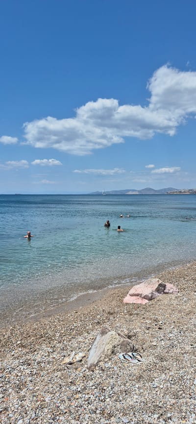 A wide shot captures a tranquil beach scene in Paleo Faliro, Greece. The foreground is a pebble beach with scattered rocks and a pair of white sandals and flip-flops. The clear, shallow water laps gently at the shore. Several people are visible enjoying the water, some swimming while others are partially submerged. In the middle ground, a sailboat glides across the calm sea. The background features a hazy coastline with hills and buildings, under a bright blue sky dotted with fluffy white clouds. A distant cargo ship is also visible on the horizon. The overall impression is one of a peaceful summer day at the seaside.