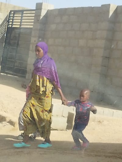 This image captures an adult female and a young child walking on a dirt path in Mariri, Nigeria, during daylight. The adult female, positioned on the left, wears a purple headscarf, a yellow and green patterned dress, and light blue flat shoes. Her left hand holds the right hand of the young child, who walks slightly ahead. The child, with a shaven head, is dressed in a dark patterned long-sleeved shirt, dark trousers, and pink shoes. Partially visible behind the adult female are the lower legs of a third individual. The background features an unplastered grey cinder block wall extending from the center-right, and a dark metal gate with horizontal bars on the left. The ground is a light brown, uneven dirt surface.
