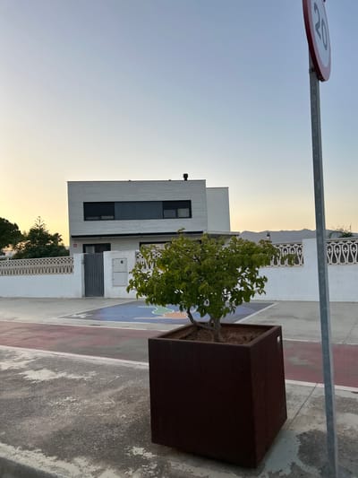 The image depicts a residential scene in Grao, Spain, during twilight. In the foreground, a square, dark brown planter contains a small, leafy green tree. To the right, a metal pole supporting a circular speed limit sign (indicated by the number "20") stands vertically, partially obscuring the sky. Behind the tree and planter, a modern, two-story white house with dark-rimmed windows is visible. The house is bordered by a white decorative fence. Adjacent to the house, a gray metal gate is present. The ground in front of the house appears to be a court with colorful markings, possibly a playground or sports area. The sky transitions from a pale yellow near the horizon to a light blue at the top, suggesting either sunrise or sunset. The overall environment appears to be a quiet, suburban setting.