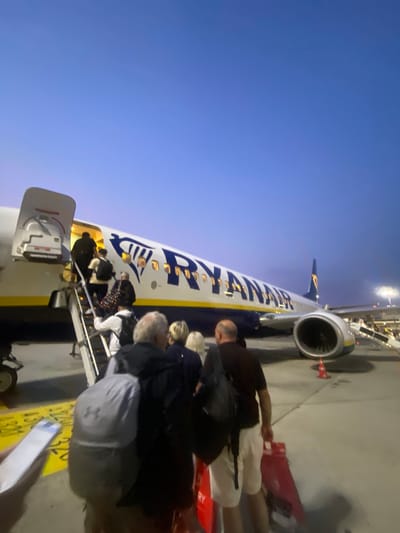 In Ferno, Italy, passengers are boarding a Ryanair airplane via a mobile staircase at dusk. The sky is a deep blue, transitioning to lighter hues near the horizon. The aircraft, emblazoned with "RYANAIR" in large blue letters and the distinctive yellow and blue logo, is parked on the tarmac. Several individuals are visible on the staircase and on the ground, carrying luggage. The scene suggests an airport environment with runway markings and lighting visible in the background.