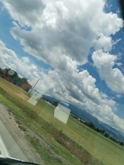 The image depicts a landscape scene from a moving vehicle in Kazichene, Bulgaria. The foreground shows a portion of a road and a grassy embankment with some gravel. The middle ground features a vast, green field, possibly wheat or barley, with some taller green grass along the edge. In the distance, there are trees, a power line pole with wires, and some buildings. Beyond that, a range of mountains can be seen under a sky filled with dramatic, fluffy white and grey clouds against a bright blue sky. The image appears to be taken through a window, as evidenced by reflections on the surface.