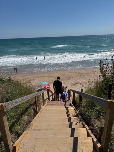 A wide-angle shot captures a sunny day at a beach in Shabla, Bulgaria, with a wooden staircase leading down to the sand. A man, wearing a black t-shirt and dark shorts, walks down the stairs carrying a blue cooler. At the bottom of the stairs, a pink flamingo float and a blue umbrella are placed on the sand. In the distance, the turquoise sea meets the sandy shore, with several people enjoying the water. The sky is clear and blue.