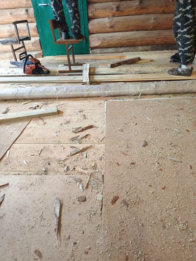 The image depicts the interior of a wooden structure, likely a cabin or workshop, in Islambaevo, Russia. The scene appears to be in a state of renovation or construction, with visible wooden planks, sawdust, and debris scattered across the floor.
In the background, a green door is set into a log wall. To the left of the door, a person's legs, clad in camouflage pants and dark shoes, are visible standing on a metal stool. A chainsaw rests on wooden planks in front of the person. To the right of the door, another person's legs, also in camouflage pants and distinctive patterned footwear, are visible standing on a wooden floor.
The foreground is dominated by the floor, which is covered in light brown particle board and numerous wood chips. Several wooden planks and fragments are scattered across the floor, suggesting active woodworking or demolition. A rolled-up plastic sheeting is also visible along the edge of the floor. The overall impression is one of a functional, albeit somewhat untidy, work area.
