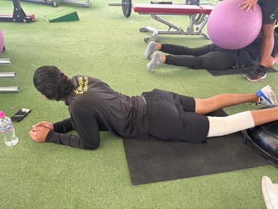 In a gymnasium, two individuals engage in prone exercises on black mats atop artificial turf. The person closer to the viewer is lying on their stomach with their elbows bent, resting on their forearms. The lower part of one leg is wrapped in white bandages. Adjacent to them is a water bottle and a phone. Behind them, another person lies prone, with a purple exercise ball placed on their lower back, and an individual assists in stabilizing the ball. Weightlifting equipment surrounds them.