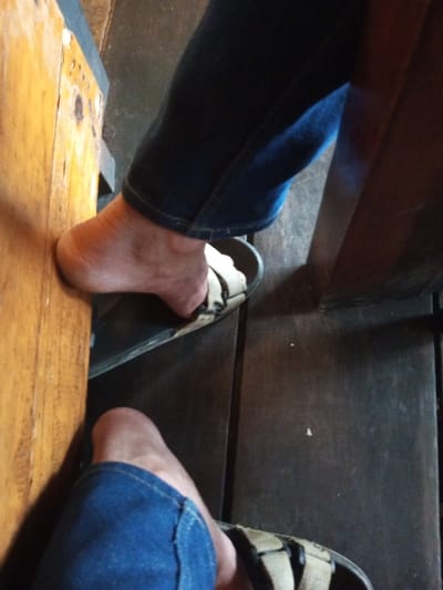 A close-up perspective reveals two feet adorned in denim jeans and tan sandals. The individual's bare heels and toes are visible, peeking out from beneath the fabric and straps. One foot is positioned next to a wooden table leg, while the other rests further back on a dark, wooden surface. The scene suggests a casual, relaxed setting, potentially in a rustic or informal environment.