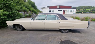 A vintage, cream-colored Cadillac Fleetwood Brougham is parked on a paved lot in Ørsta, Norway. The car, seen from its right side, has a dark brown vinyl roof and chrome trim. In the background, a white and red building with a dark roof is visible, with an air conditioning unit mounted on the upper wall. Green foliage and rocks are to the left of the car, and a gently sloping, treed hillside with scattered houses is visible further in the distance. The sky is overcast.