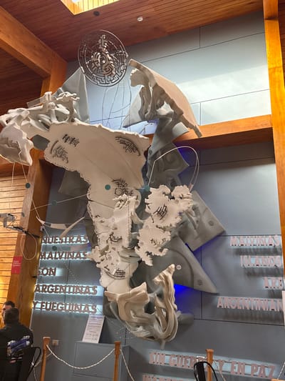 The image depicts an indoor scene in Tolhuin, Argentina, showcasing a large, multi-component art installation mounted on a light grey-blue wall. The installation features numerous irregularly shaped, textured, light-colored (possibly off-white or light grey) pieces, some resembling fragmented landmasses or maps, with specific areas marked by numbers (e.g., 52, 103) and abstract patterns. These pieces are layered over darker grey, more geometric and angular background forms.
Suspended from the ceiling above the installation is a circular metallic art piece, designed as a complex labyrinth or maze. Thin wires or lines, intermittently punctuated by small turquoise spheres, connect various elements of the map-like structures. Transparent acrylic or glass elements are also integrated into the design, appearing to curve around parts of the artwork.
To the left of the installation, white three-dimensional block letters vertically spell "NUESTRAS MALVINAS SON ARGENTINAS Y FUEGUINAS." Correspondingly, on the right side of the installation, similar block letters translate this sentiment into English, reading "MALVINAS ISLANDS ARE ARGENTINIAN." Further below on the right, additional text states "SIN GUERRA Y EN PAZ." Blue accent lighting illuminates specific sections of the darker background elements within the installation.
The environment features a high, wood-paneled ceiling with visible skylights. Large, exposed wooden structural beams (columns and horizontal members) frame the wall and the installation, suggesting a modern architectural interior. A low barrier, consisting of wooden posts connected by a white braided rope, demarcates the viewing area at the bottom of the frame. Behind this barrier, a small white information plaque with text and icons is visible.
Two individuals are partially visible in the lower-left corner of the frame, positioned behind the rope barrier. One person, viewed from the back and side, wears a dark shirt with white text. A second person's head is partially visible beside them. Both appear to be static, observing the exhibit or present in the space. No direct interactions between the individuals or with the exhibit are discernible.