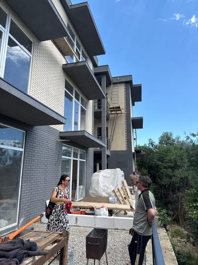 In the city of Yalta, Ukraine, two men and one woman are standing on a balcony of a multi-story building under construction. The building features a light-colored brick facade with dark grey accents and cantilevered balconies. The woman, wearing a black and white patterned dress, stands to the left, facing away from the camera. She is holding a red object and a bag over her shoulder. The two men are positioned to the right, looking towards the building. One man, wearing a grey and green polo shirt and black pants, has his back to the camera. The other man, wearing a light-colored shirt, is partially visible between them. In the foreground, construction materials such as wooden pallets and buckets are visible, along with a portable grill. The background is dominated by lush green trees and a clear blue sky.