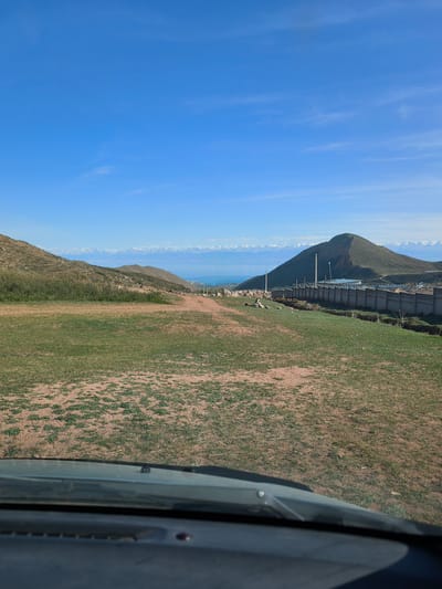 The image captures a vast outdoor landscape, likely taken from inside a vehicle, with the dashboard and windshield visible at the bottom. The scene features rolling hills, a large body of water, and distant snow-capped mountains under a clear, bright blue sky with only a few faint cirrus clouds.
In the immediate foreground, the ground is a mix of sparse green grass and bare reddish-brown earth, suggesting dry conditions. On the left, a grassy hill slopes upwards. To the right, a long, grey concrete fence topped with barbed wire stretches across the middle ground, following the contours of the land. Utility poles are visible along and behind the fence. Behind the fence, a prominent, green, conical-shaped hill rises. Partially visible beyond the fence and conical hill are some buildings.
In the distance, a large expanse of deep blue water, likely Lake Issyk-Kul, stretches across the horizon. Beyond the lake, a majestic range of snow-capped mountains, characteristic of the Tian Shan range, forms a striking backdrop under the clear sky.
The image depicts a bright, sunny day. No people or specific activities are observable, though the presence of the vehicle suggests travel or a stop to admire the view. There is no visible text in the scene. The overall setting is a rural or semi-rural mountainous region, consistent with the provided location of Cholpon Ata, Kyrgyzstan.