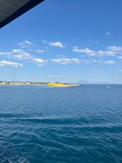 The image depicts a wide-angle view of a body of water, likely the Ionian Sea, with a yellow ferry docked at a harbor on the left. The ferry is branded with "Levante Ferries" and the number "11380". Behind the ferry, a coastal town with buildings and a breakwater extends along the shoreline. In the distance, hazy mountains rise against a bright blue sky dotted with white clouds. A small white boat is visible in the middle of the water, further out to sea. The foreground shows the textured surface of the water, with white foam trails suggesting movement from a vessel. The top left corner of the image is partially obscured by a dark blue structure, likely part of a larger boat or structure from which the photo was taken. The location is the Municipal Unit of Kastro - Kyllini, Greece, a port town known for its ferry connections to the Ionian Islands.