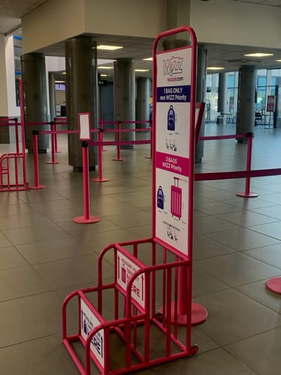 The image shows the interior of an airport terminal in Pyrzowice, Poland, with a sign displaying Wizz Air baggage restrictions. There is a pink metal frame with a sign that says "Measure your trolley bag here" and a red rope barrier. The rope barrier is made of a metal post with red rope attached to it. The image shows a single large sign with information regarding baggage restrictions. The sign says "Wizz Wizzair.com 1 bag only non Wizz Priority 2 bags Wizz Priority." The sign features images of a backpack and a suitcase. The location is inside the airport, which is indicated by the presence of a rope barrier, sign, and airport terminal. The floor is tiled and there are no people present in the image. The image captures the moment of a passenger checking baggage regulations before check-in. The image shows a large sign with a diagram and a description of how to properly check baggage at the airport.