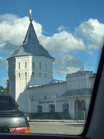 The image shows the Annunciation Cathedral of the Nikolo-Vyazhishchsky Monastery, located in the city of Dzerzhinsky, Russia. The cathedral is a prominent white stone structure with a distinctive octagonal tower topped with a golden angel. The tower has several rows of arched windows. Adjacent to the tower is a lower, white stone building with a serrated parapet and arched entrances. The facade of this building features a sign with Cyrillic lettering that reads "Монастырская кофейня" (Monastery Coffee House). In front of the coffee house, there are metal barriers and some signage. The scene is viewed from inside a vehicle, with the reflection of the car's interior visible in the foreground. A black car is parked on the left side of the frame. The sky is blue with scattered white clouds.