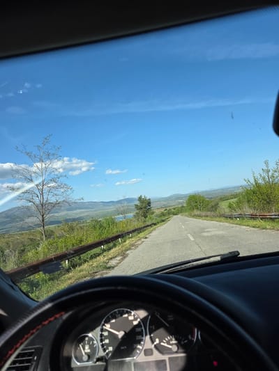The image depicts a view from inside a car, looking out onto a road in Lelintsi, Bulgaria. The road is asphalt, with a guardrail on the right side. Beyond the guardrail, there is a landscape of green hills and vegetation. In the distance, a body of water is visible. The sky is blue with scattered white clouds. The car's dashboard and steering wheel are partially visible in the foreground. The speedometer reads approximately 100 km/h.