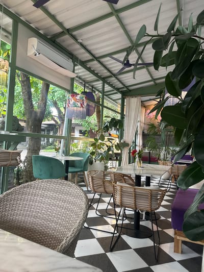 This image shows an interior scene, likely a restaurant or cafe in Celuk, Indonesia. The space has a greenhouse-like structure with a white ceiling, green metal framework, and large windows. Outside the windows, lush green trees and foliage are visible. Inside, a table with a white marble top is surrounded by wicker and plush chairs. The floor is covered in a black and white checkered pattern. A ceiling fan is visible in the upper left of the frame. The scene is well-lit with natural light streaming through the windows.