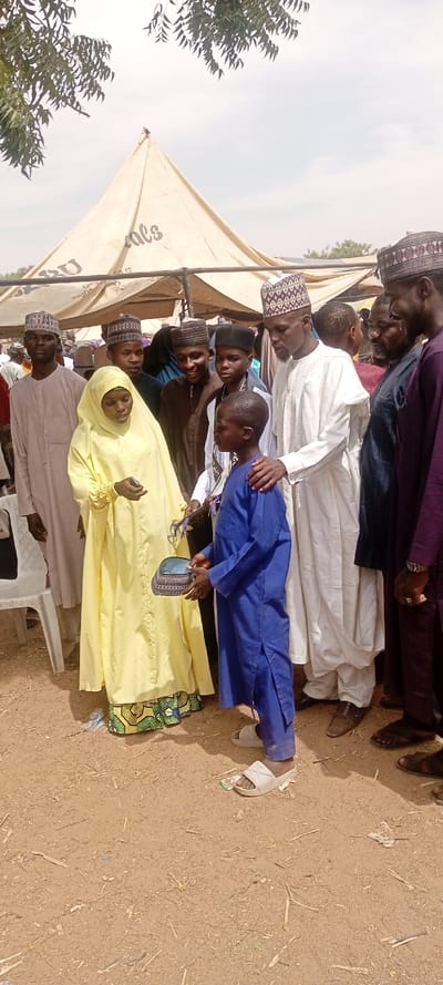 The image depicts an outdoor gathering in Dorayi, Nigeria, featuring nine individuals under a light-colored canvas tent with visible dark lettering. In the foreground, a young boy in a blue traditional outfit and white slide sandals stands centered, holding a patterned cap or small basket in both hands, facing a woman in a bright yellow full-length dress with a patterned hem. The woman holds a small dark object in her right hand and looks towards the boy. To the boy's right, an adult male in a white traditional robe and patterned cap places his right hand on the boy's shoulder. To the woman's left, a man in a light beige robe and patterned cap stands. Additional males in various traditional garments and caps are visible behind and to the right of the central figures, some smiling. The ground is unpaved, light brown dirt or sand, scattered with small pieces of wood and debris. Green foliage from trees is visible at the top corners against a light, possibly overcast, sky. A white plastic chair is partially visible on the far left.