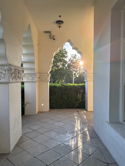 The photograph captures a sunlit walkway with distinct architectural features. The walkway is paved with square tiles, laid diagonally. On the left, a white column with ornate carvings supports an archway. Further down the walkway, another similar archway is visible, framing a dense green hedge and trees beyond. Sunlight streams through this arch, creating bright reflections on the tiled floor. Above the archway, a light fixture and a security camera are mounted on the ceiling. The overall impression is one of an outdoor corridor with classical architectural influences, bathed in the warm light of the sun. The image does not contain any identifiable people or specific landmarks that would pinpoint a precise location within Glendale, United States of America, beyond it being an outdoor area.