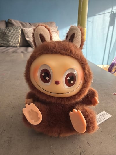 A brown, plush toy with a large plastic face lies on a gray surface. The toy has fuzzy fur around its face and ears. The face is light tan with large brown eyes. The toy is lying on its back, with its plastic feet pointed upwards. In the background, there is a glimpse of a blue wall, a gray couch, and a window with blinds. The image was taken indoors, likely in a private residence. There is no information about this image that suggests it was taken in the city of Lørenskog, Norway.