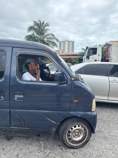 The image shows a woman in the driver's seat of a dark blue minivan. She is wearing a white t-shirt and a green and white patterned headscarf. She has earrings and appears to be putting something to her mouth with her right hand. Another person, a man wearing a grey baseball cap and a dark t-shirt, is visible in the passenger seat behind her.
The minivan is parked on a paved road, with a white truck and a white car behind it. A tall building and palm trees are visible in the background, suggesting an urban setting in a tropical or sub-tropical location. The sky is overcast, indicating it might be a cloudy day. The overall scene appears to be a street in Ikoyi, Nigeria. The van has visible wear and tear, with scratches and rust on the exterior. The wheel of the minivan is also visible, appearing somewhat dirty. There is no discernible text in the image.