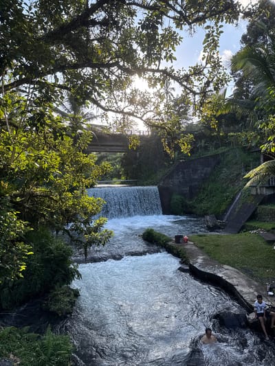 This image depicts a river with a cascading waterfall in Rendang, Indonesia. The sun shines brightly through the leaves of a large tree in the foreground, casting dappled light on the water and surrounding vegetation. A concrete bridge spans the river in the background. On the right bank of the river, a grassy area leads to a concrete walkway. Two young boys are seated on the walkway, while a man bathes in the river. There are some rocks and plants along the riverbank.