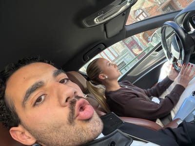 A selfie-style photograph captures two individuals inside the cabin of a vehicle in Uccle - Ukkel, Belgium. In the foreground, positioned to the left, an adult male with dark, curly hair, a mustache, and a beard is looking directly at the camera with puckered lips. He is wearing a dark jacket over a white garment.
Behind and to his right, an adult female with blonde hair tied in a ponytail occupies the passenger seat. She wears a dark brown long-sleeved top and light-colored lower garments. Her fingernails are painted red, and she has a small, round pearl-like earring in her visible left ear. Her gaze is directed upwards and forward, out of the vehicle. Her hands rest in her lap. A light blue water bottle is visible next to her left leg, and a dark electronic device is near her right leg.
The vehicle's interior features a dark headliner and dashboard, with brown leather upholstery on the seats. A black steering wheel with silver accents is partially visible on the left side of the car. Through the windshield, the exterior environment reveals sunlit, multi-story brick buildings with arched windows and classical European architectural details, indicating an urban setting.