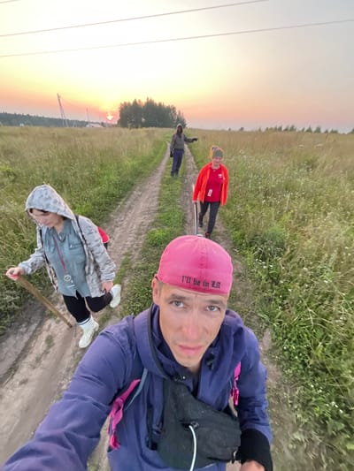 The image depicts a group of three people walking on a dirt road in a rural setting, possibly the city of Badashka, Russia, during sunset. The sky is illuminated with hues of orange and yellow, suggesting the sun is low on the horizon. The road is flanked by tall green grass and vegetation.

In the foreground, a man wearing a purple hooded jacket and a pink baseball cap is taking a selfie, with his face close to the camera. The cap has the text "I'll be in LA" embroidered on it.

Further down the road, a woman in a camouflage hooded jacket, black leggings, and white boots is walking, holding a wooden stick. She has a red backpack strapped to her.

Behind her, another woman in a bright red jacket and dark leggings is walking. She is holding a long, thin object, possibly a walking stick or a pole. A third individual, partially visible in the background, is also walking on the road.

The overall scene suggests a group engaged in outdoor activity, possibly hiking or walking, in a natural environment as dusk settles. Power lines are visible in the distance.