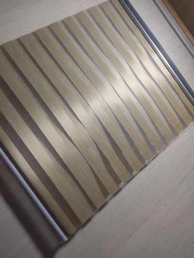 The image shows a close-up, overhead view of a window blind with a striped, wood-grain pattern. The blind is composed of alternating opaque and translucent stripes, creating a layered effect. The stripes run diagonally from the bottom left to the top right of the frame. The background appears to be a light-colored wooden floor. There are no people or specific city landmarks visible in the image.