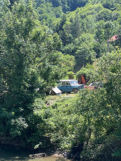 This image depicts a scene in Moračko Trebaljevo, Montenegro, characterized by dense green foliage and a body of water in the foreground. A vintage blue SUV is parked on an incline amidst overgrown vegetation. Behind the SUV, an orange piece of construction equipment, possibly an excavator, is visible. In the distance, a red-roofed house is nestled among trees on a hillside. The water in the foreground appears shallow, with debris floating on its surface. The overall impression is one of a rural, somewhat neglected setting.