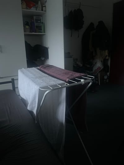 The image depicts a cluttered room, likely a bedroom or studio apartment in Hilversum, Netherlands. In the foreground, a metal drying rack holds two large towels, one light grey and the other a dark purple or maroon. These towels are neatly folded. To the left of the drying rack, a dark grey mattress is visible, covered with a duvet or sheets.
Behind the drying rack, a white closet is partially open, revealing shelves stocked with various items, including bottles and containers, suggesting toiletries or cleaning supplies. The shelves are organized with plastic organizers. More items are visible deeper within the closet, including dark clothing or bags.
To the right of the closet, a second, darker area of the room is visible. This area appears to be a wardrobe or another storage space. Several dark jackets or coats are hung on a rail, some with hoods. Bags and other items are piled on the floor and shelves in this area. A large white shopping bag with red lettering is prominent. Bottles are also visible on the floor. The overall impression is one of a lived-in space with storage challenges. The lighting is dim, with strong shadows, suggesting natural light is limited or artificial lighting is low.